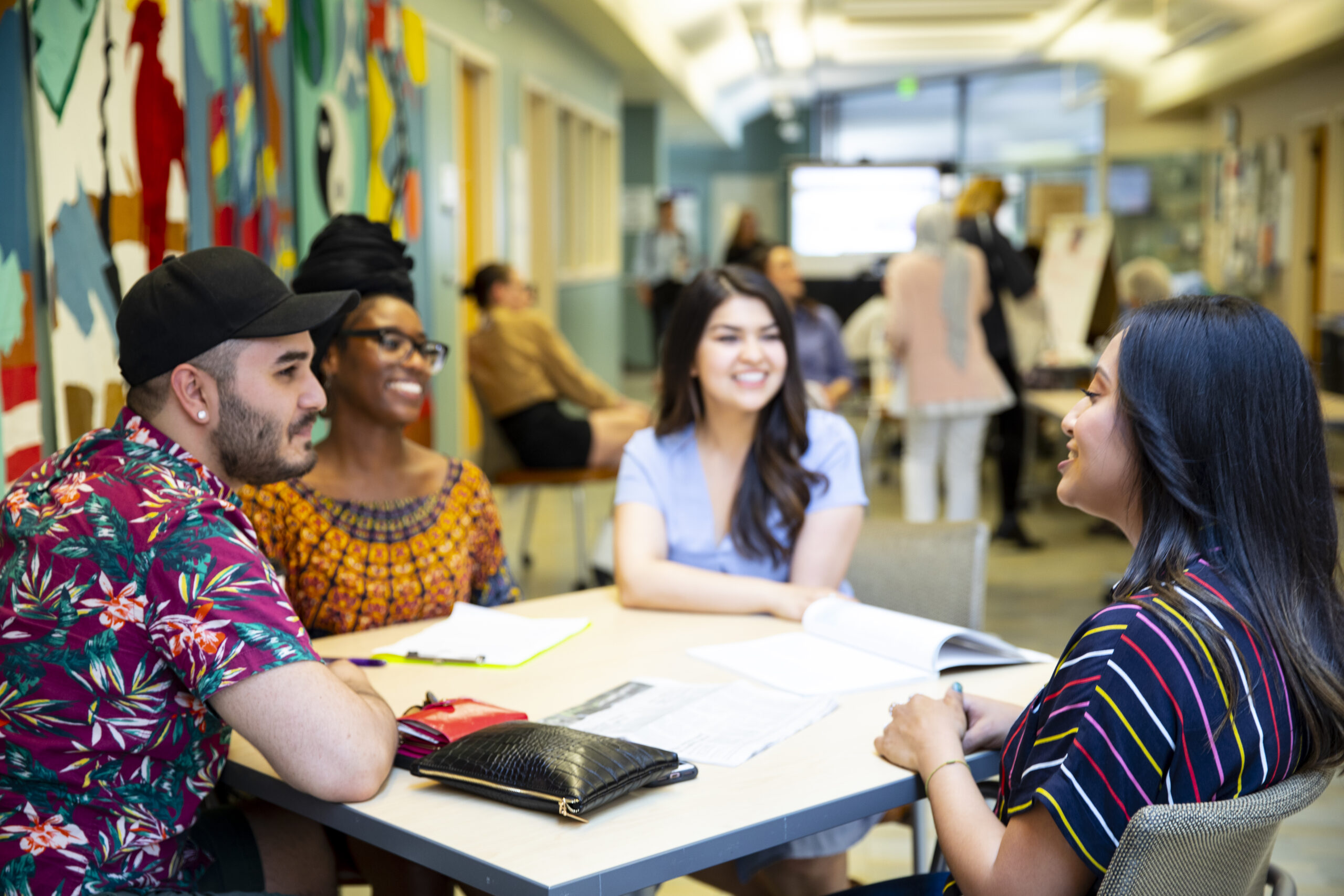 Four students sitting together at a table, smiling and talking in a brightly colored hallway