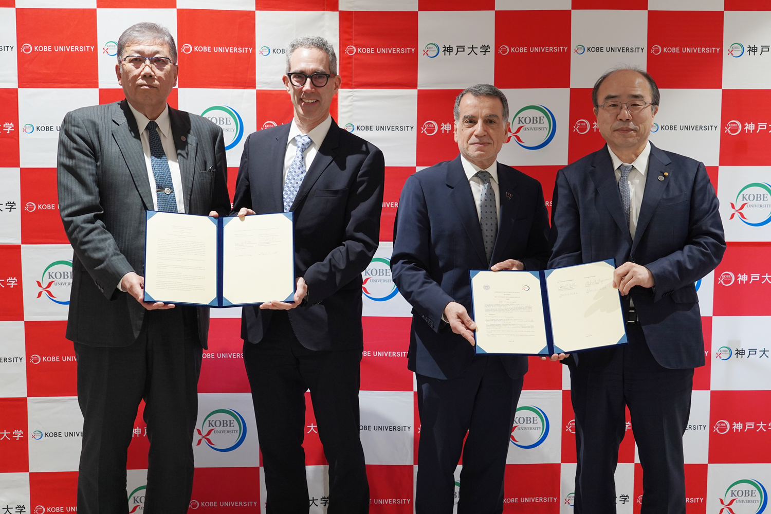 Four members from Kobe University and University of Washington stand in front of a Kobe University backdrop, holding open documents.