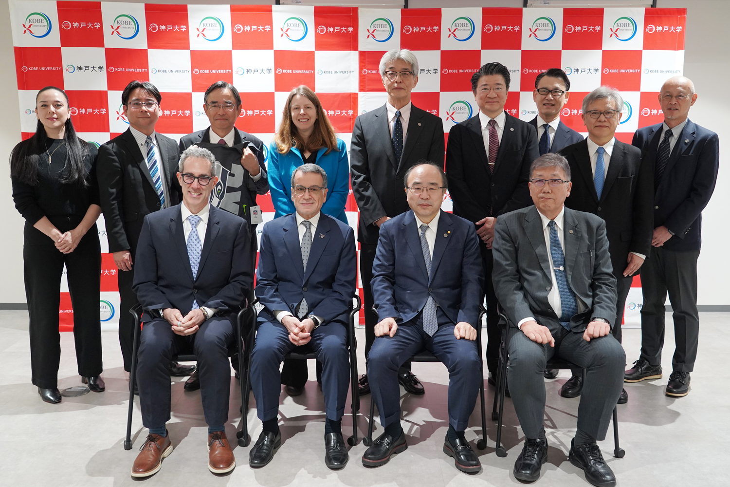 Group portrait of university officials and visitors standing and seated in front of a red-and-white Kobe University backdrop.