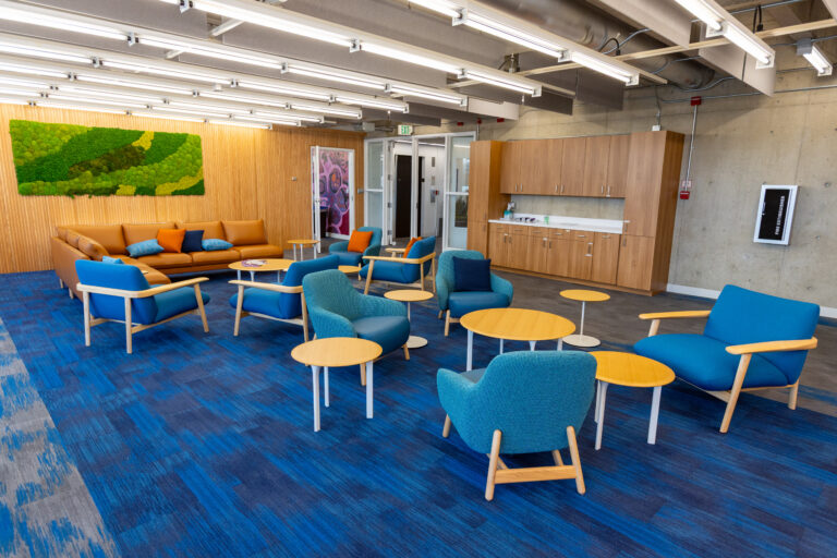 Modern lounge area with blue chairs, an orange sofa, small tables, a kitchenette, and a green moss wall installation.