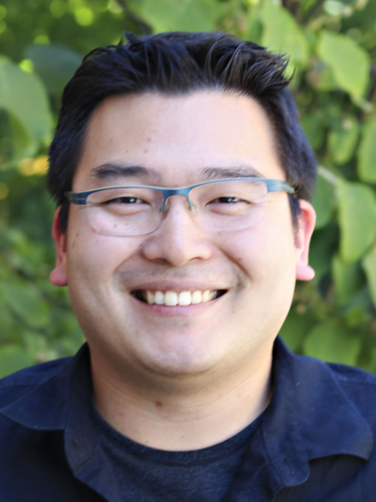 Adrian smiling outdoors, wearing glasses and a dark shirt against a leafy green background.